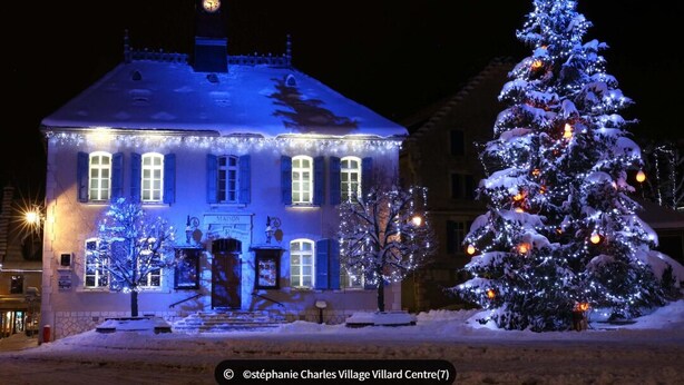 A building with a clock tower, illuminated windows, and a snow-covered Christmas tree.