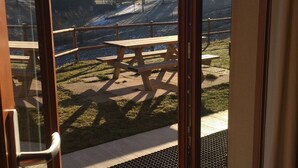 A view through a glass door of a patio area with picnic tables, a wooden deck, and a mountain backdrop.