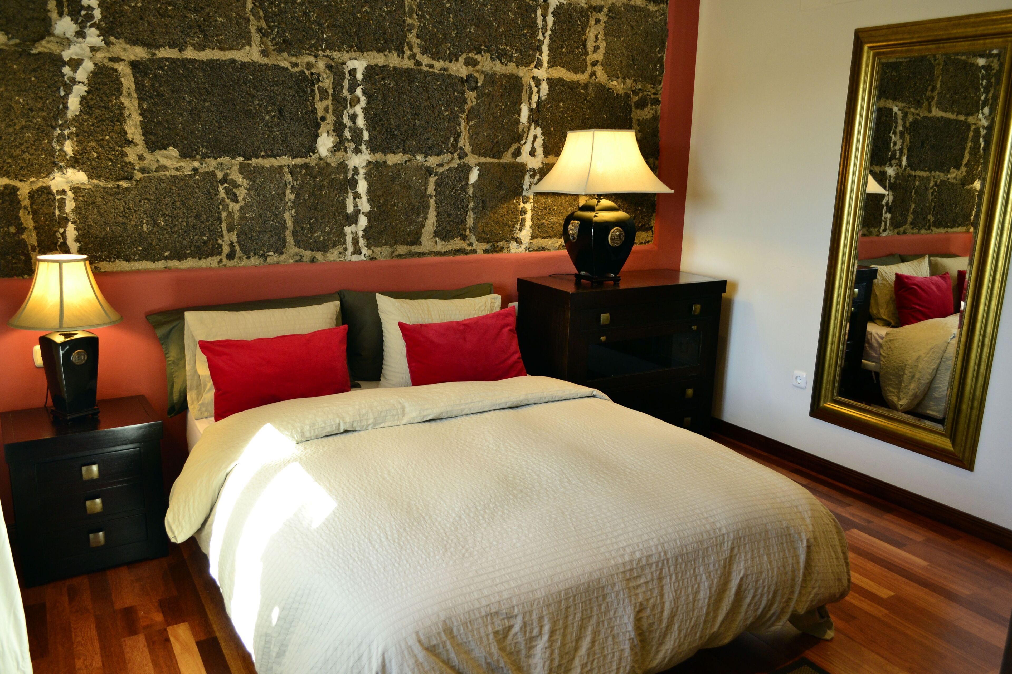 Une chambre avec un mur en pierre, un lit avec des oreillers rouges et blancs, une table de chevet avec une lampe et un miroir sur le mur.