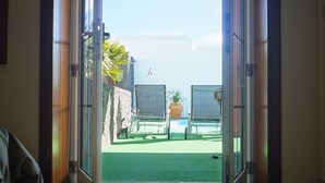 An open doorway leading to a patio with a view of the sea, a potted plant, and two lounge chairs.