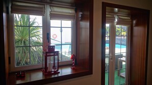 A room with a view of a pool and palm trees through large windows with blinds.