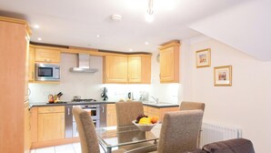 Een moderne keuken met houten kastjes, een magnetron, fornuis en een eethoek met een glazen tafel en twee stoelen.