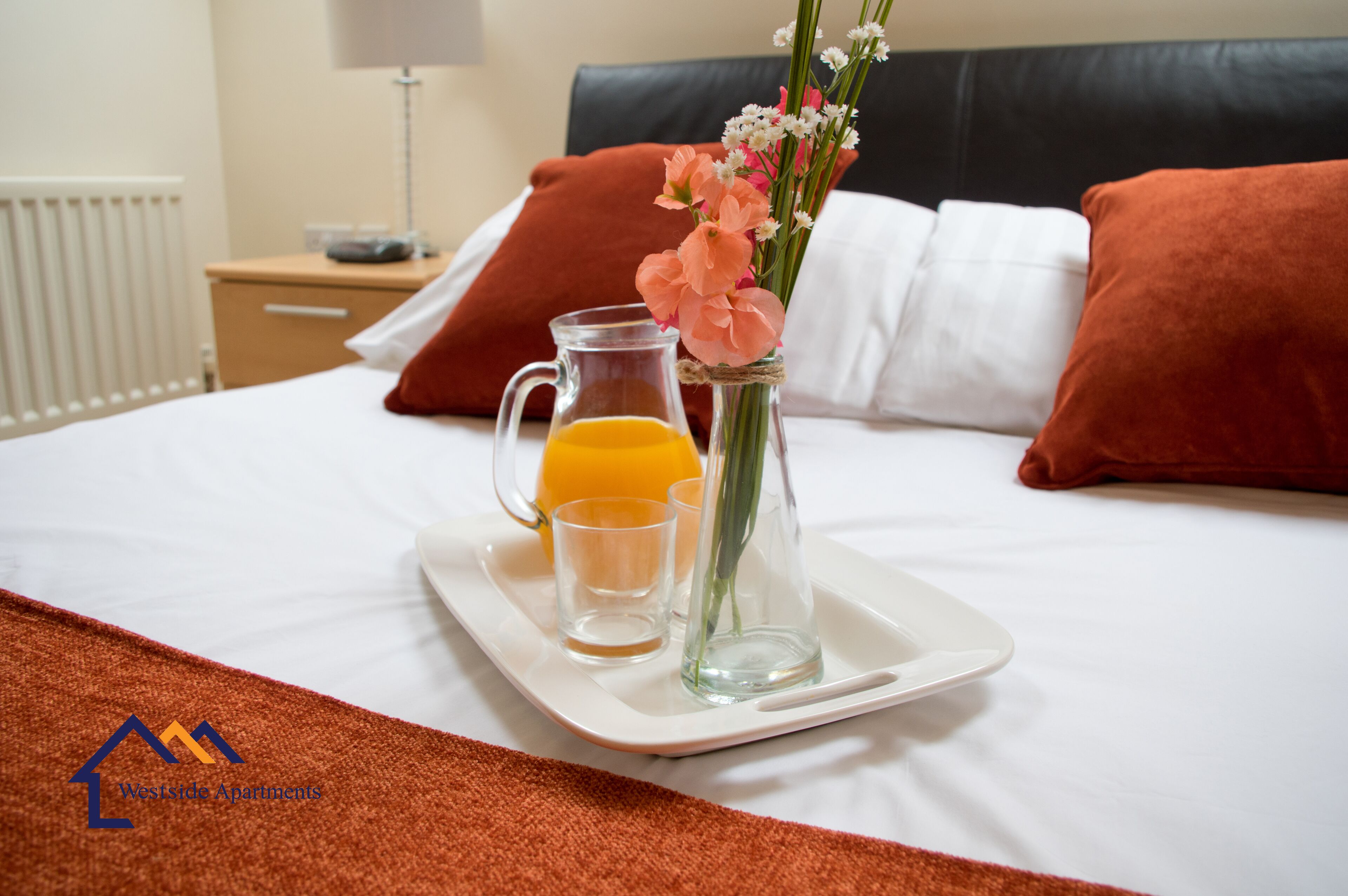 Un lit bien fait, sur lequel se trouve un plateau avec une carafe de jus d’orange, un verre et un vase rempli de fleurs.