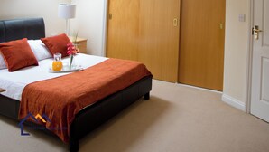 Een slaapkamer met een bed, nachtkastje, lamp, dienblad met een glas sinaasappelsap en een houten garderobe.