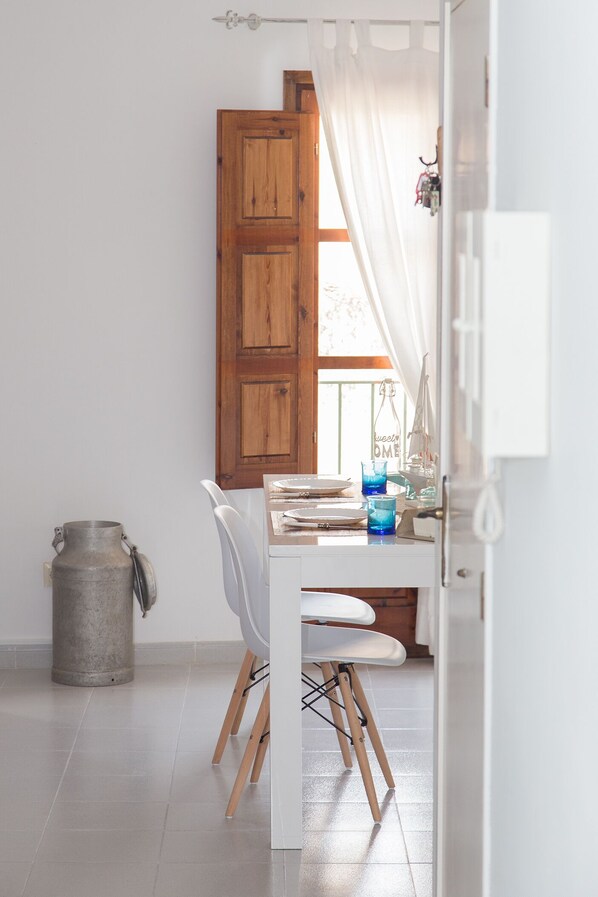 A dining area with a wooden door, white curtains, a table set with plates and glasses, and a metal container. - Very bright apartment 1 min walk from the beach of Tossa recently refurbished (Tossa de Mar)