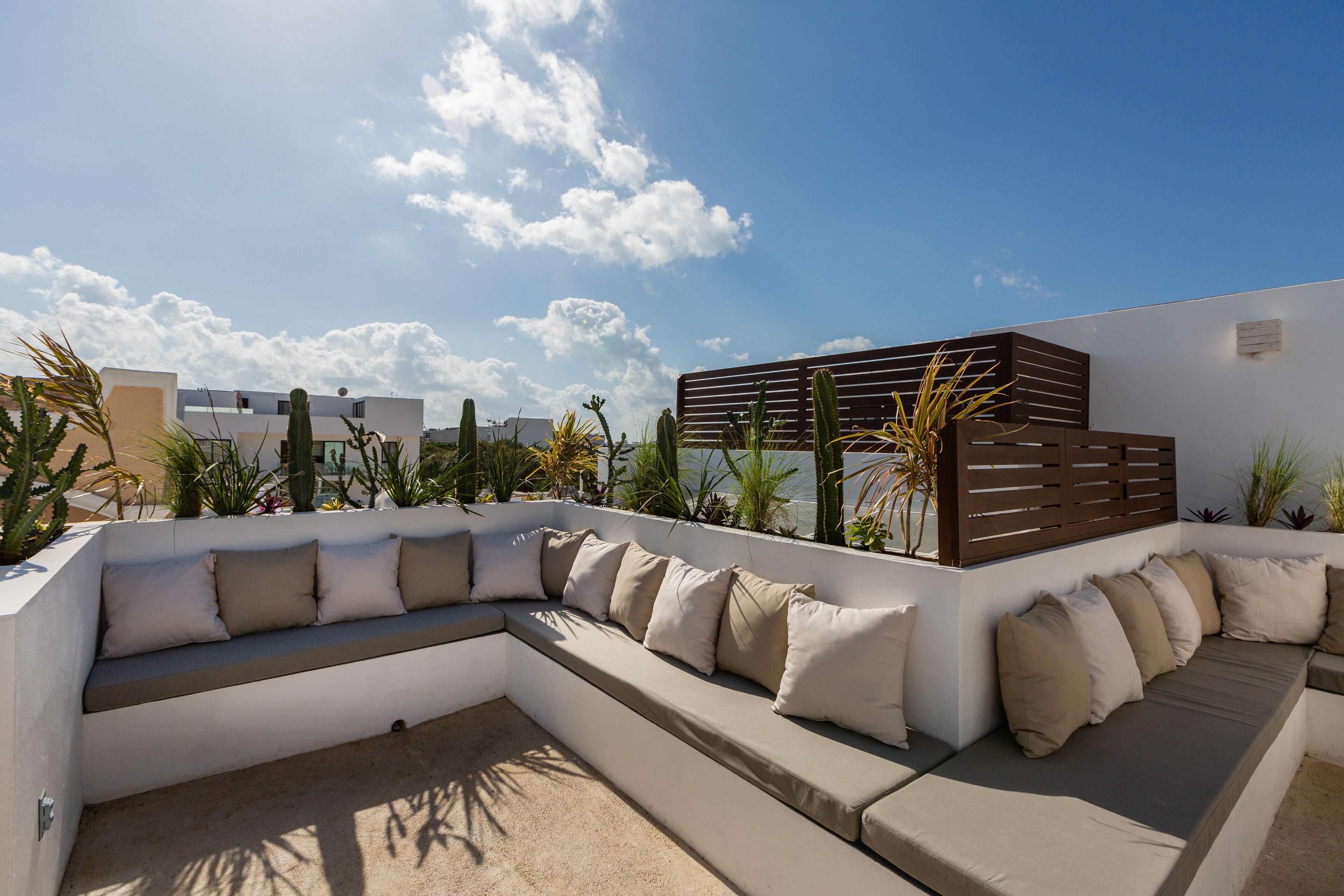 A rooftop seating area with a curved sofa and cushions, surrounded by potted plants and a clear sky.
