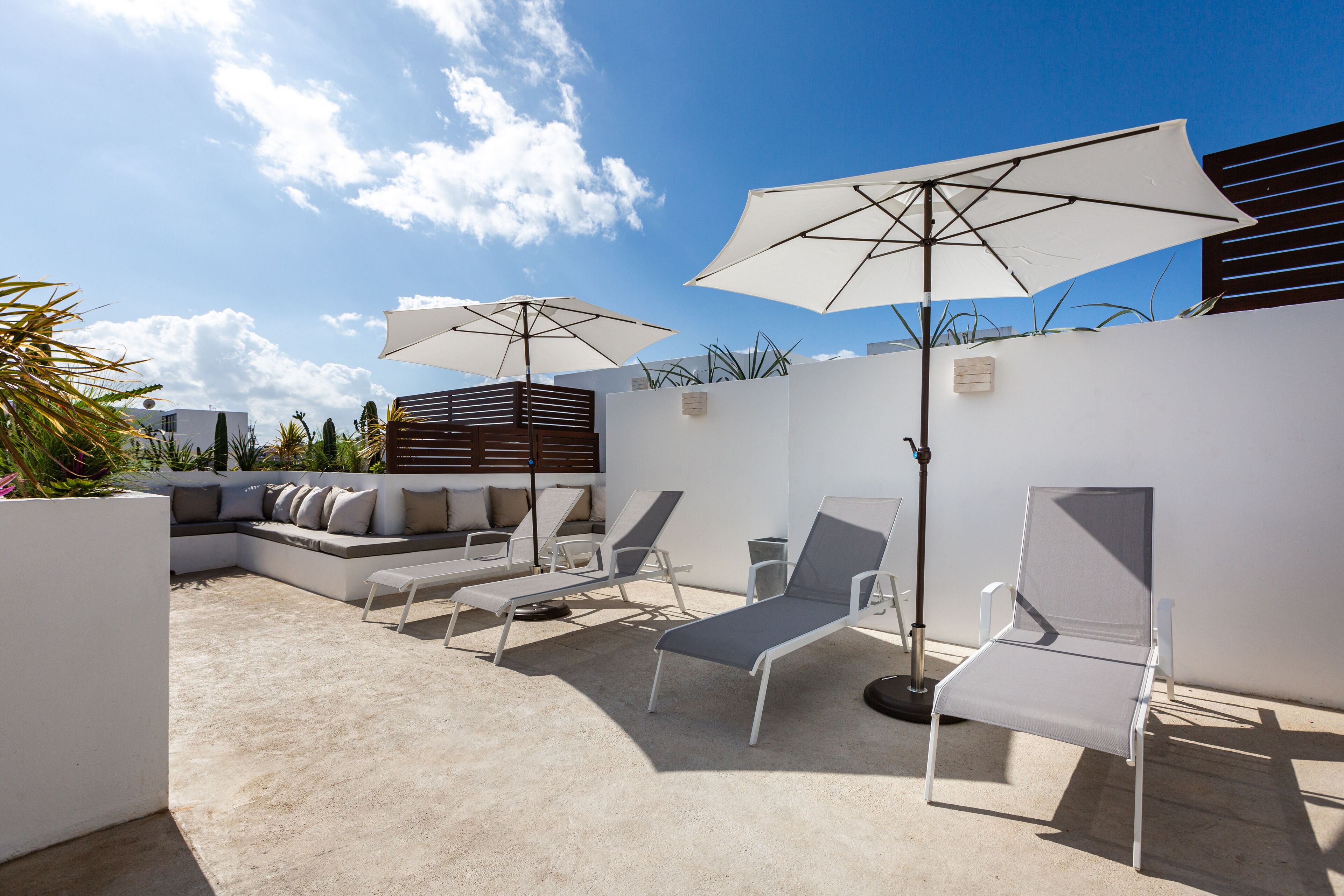 Uma área de lounge no terraço com cadeiras brancas, dois grandes guarda-sóis e um sofá moderno com almofadas.