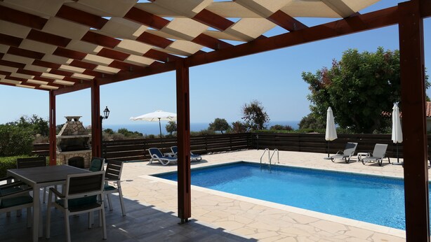 A covered outdoor pool area with a stone barbecue, lounge chairs, and a view of the sea. - Spacious,quiet villa in a secluded tranquil location with all amenities nearby (Peyia)