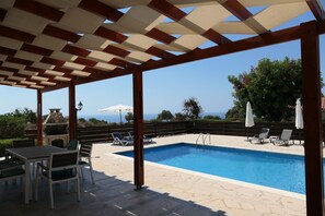 Un espace piscine extérieur abrité, doté d’un barbecue en pierre, de chaises longues et offrant une vue sur la mer.