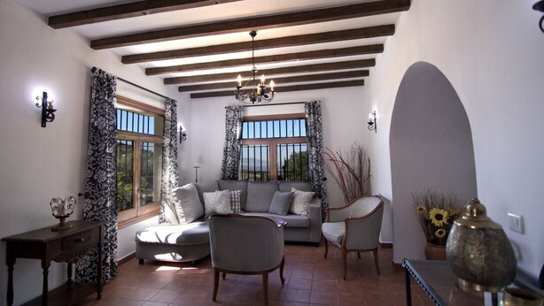 Ein Wohnzimmer mit einer Couch, Sesseln, einem Tisch und einem Chandelier. Es gibt Fenster mit Vorhängen, eine Vase mit Sonnenblumen und ein Wanddekor.