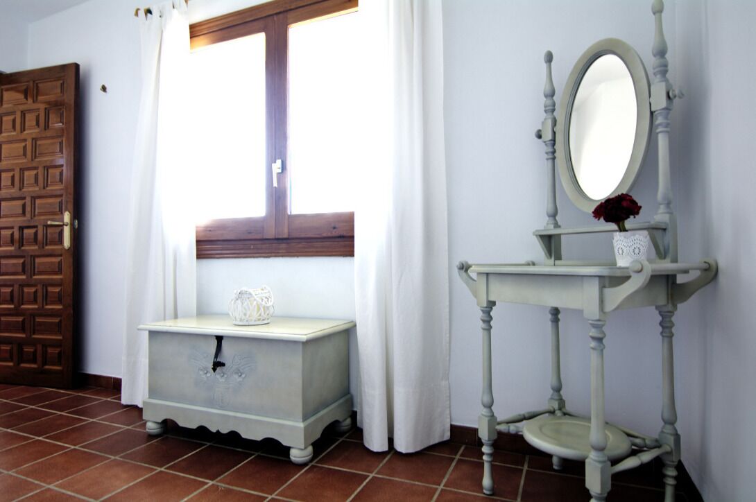 A room with a wooden door, a window with white curtains, a small white dresser with a vase, and a pedestal table with a mirror and a vase with a red flower.