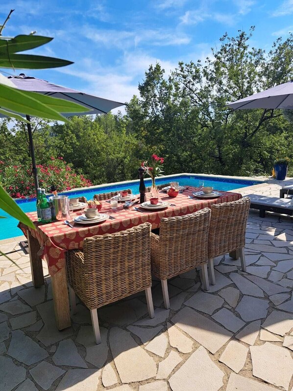 Un espace de restauration en plein air, avec des chaises en osier et une table dressée pour quatre personnes, donnant sur une piscine entourée d’une végétation luxuriante.