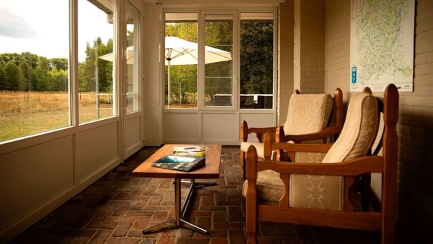 A sunroom with large windows, a wooden chair, a table with books, and a map on the wall. - Unique vacation home with a hot tub in nature (Meeuwen-Gruitrode)