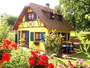 Een traditioneel vakwerkhuis met een schuin dak, een tuin met rode en roze bloemen, en een terras met tuinmeubilair.