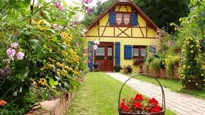 Ett traditionellt hus med korsvirkesfasad och en välskött trädgård, och i förgrunden står en metallkorg fylld med röda blommor.