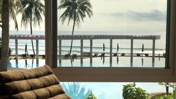 Ein Zimmer mit großem Fenster mit Blick auf einen Strand, ein Pool und eine Lounge mit gemusterten Kissen.