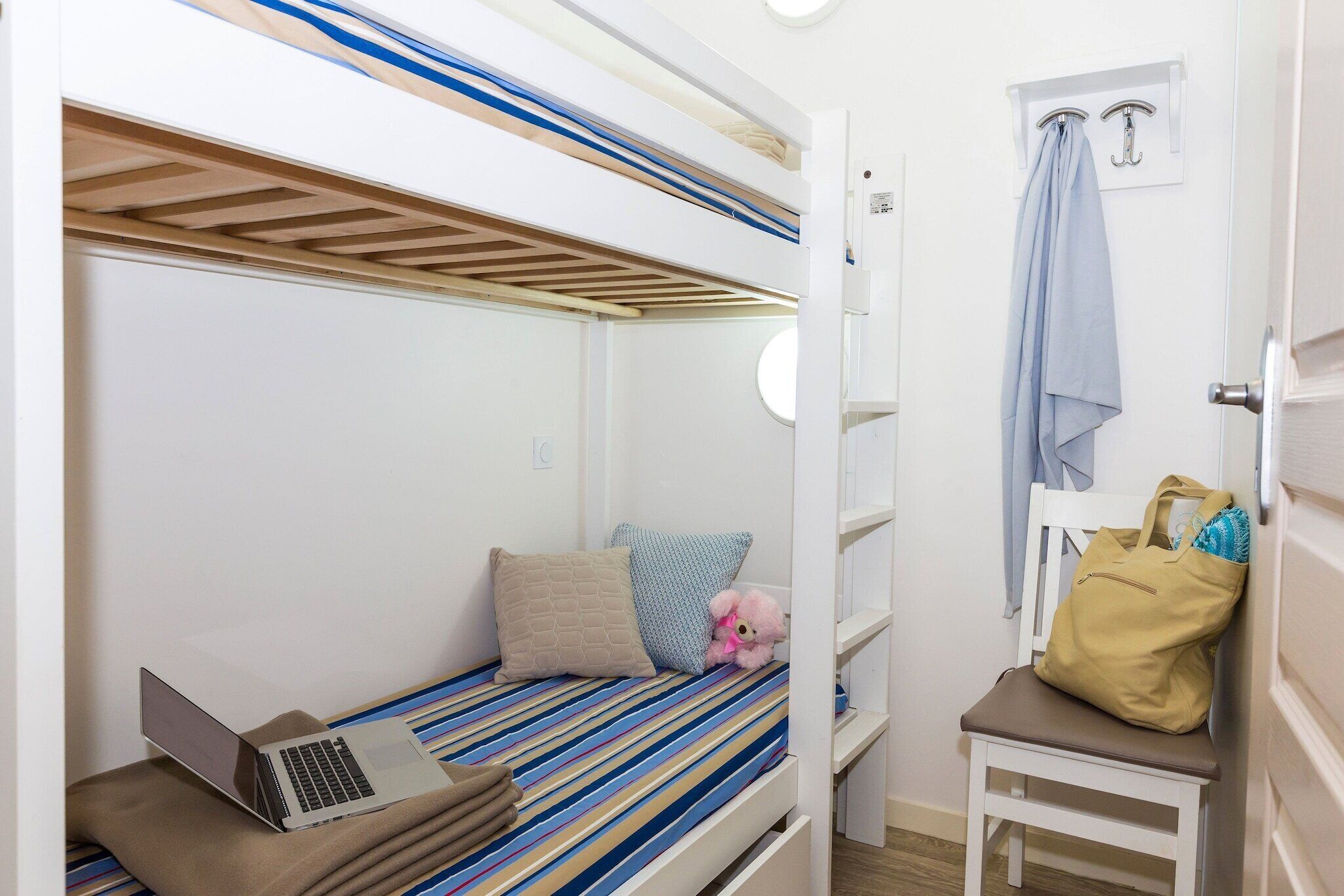 Una cama litera con un ordenador portátil en la litera inferior, un osito de peluche y un bolso en una silla.