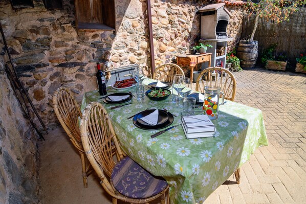 A stone-walled outdoor dining area with a table set for two, featuring a green floral tablecloth, wicker chairs, and a rustic stone oven in the background.