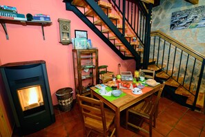A cozy dining area with a table set for two, a wood-burning stove, and a staircase with a stone wall backdrop.
