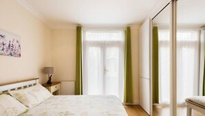 A bedroom with a bed, a nightstand, a lamp, a radiator, and a large window with curtains. - Bikki Apartments, Bikki Apartments (Harrow)