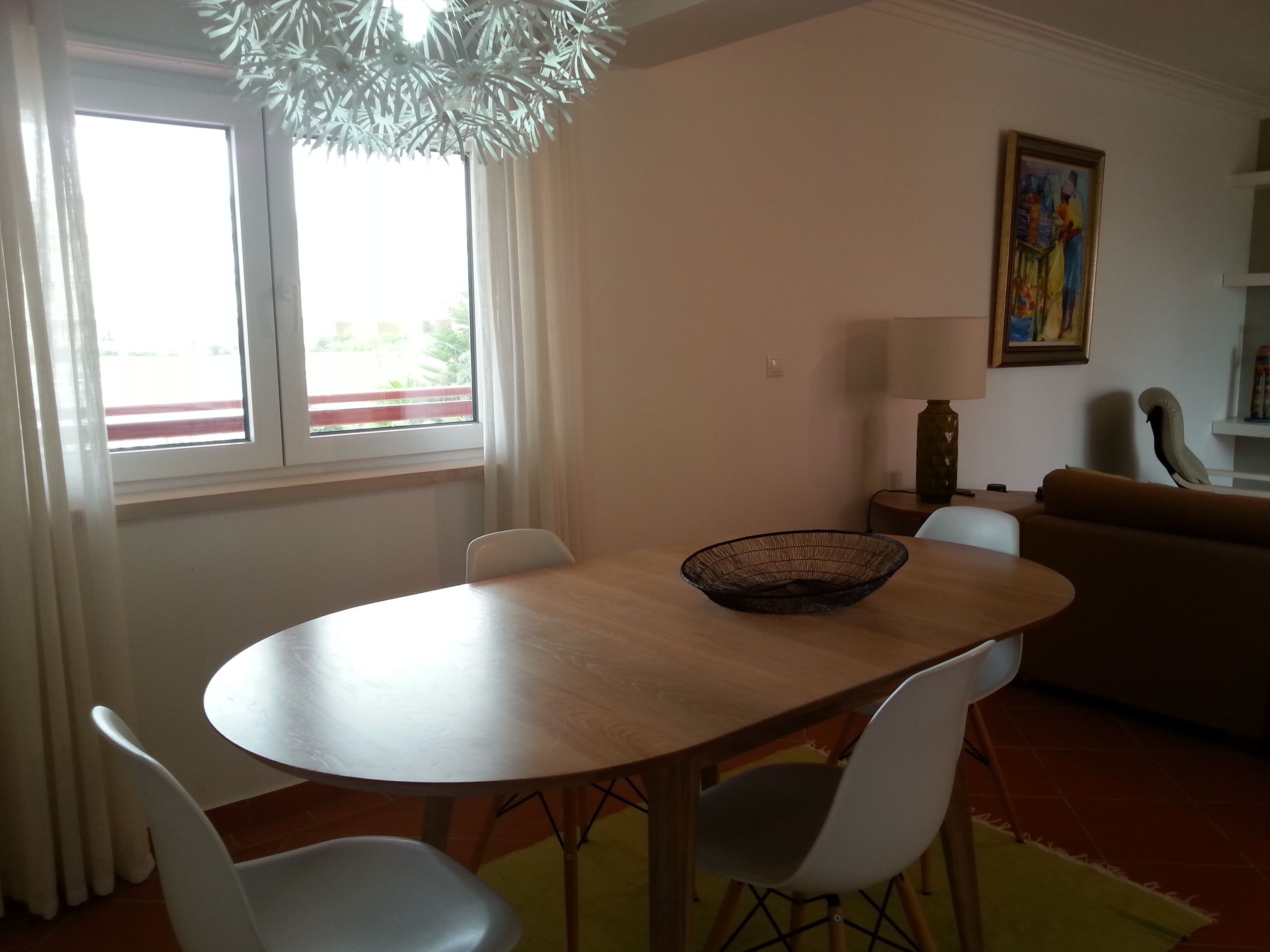 A dining area with a round table, white chairs, a black woven bowl, a window with curtains, a painting on the wall, and a sofa in the background.