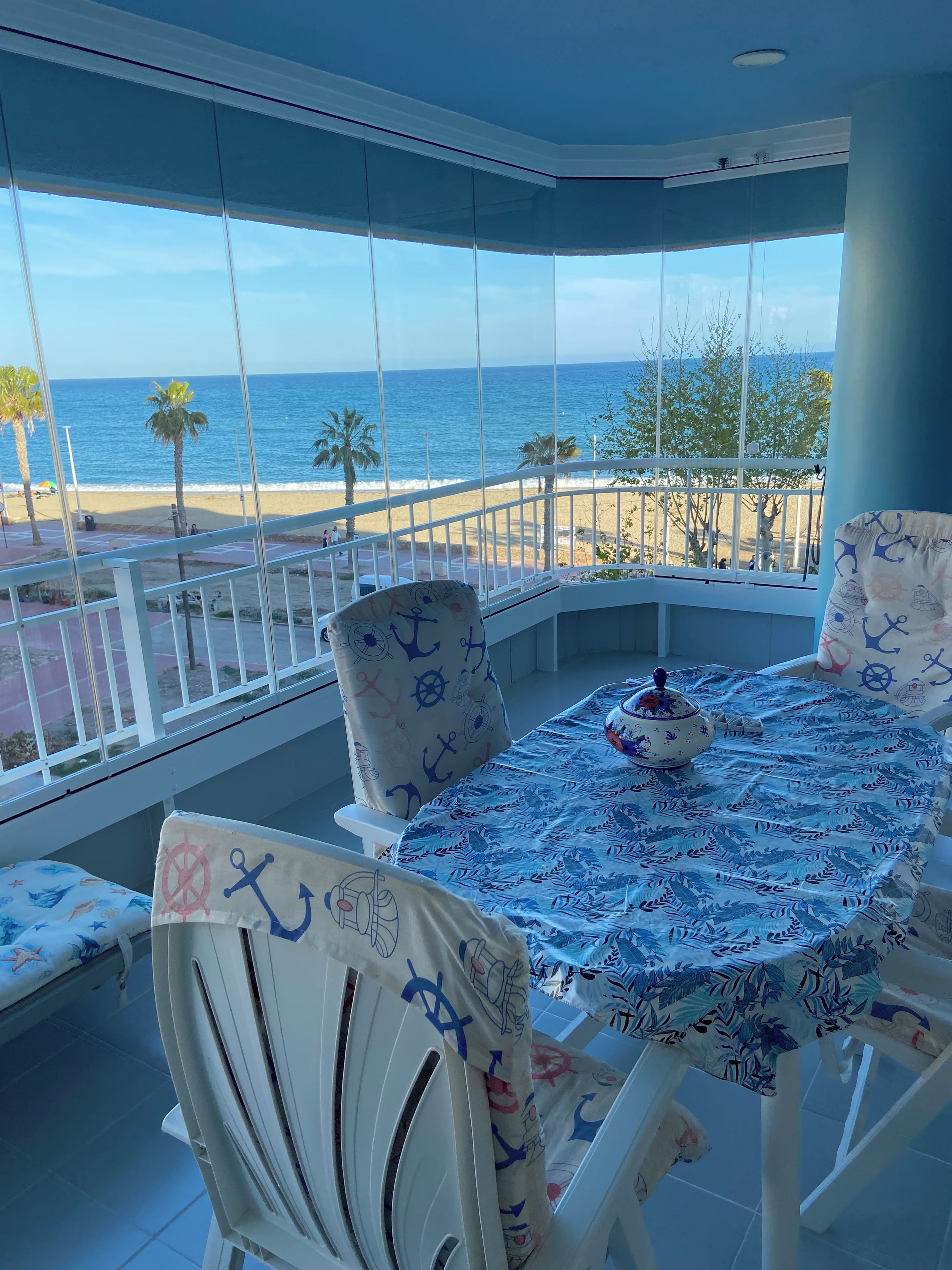 A balcony with a view of the beach and ocean, furnished with a table and chairs, and a large window.
