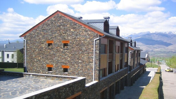 A stone building with a sloped roof and multiple windows, located next to a paved walkway with a view of distant mountains. - Cozy apartment in Alp with garden and parking, 10 minutes from the ski slopes! (Alp)