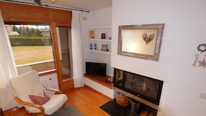 A cozy living room with a fireplace, wooden flooring, a comfortable chair, and a view of the outdoors through a large window. - Cozy apartment in Alp with garden and parking, 10 minutes from the ski slopes! (Alp)