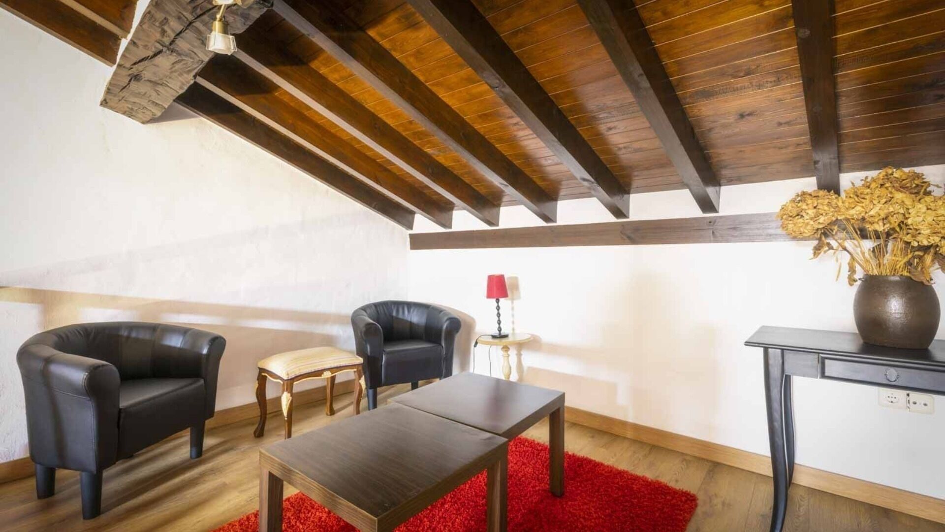 Ein Zimmer mit Holzboden, zwei schwarzen Ledersesseln, einem kleinen Holztisch, einem roten Teppich und einer Vase mit Trockenblumen auf einem Beistelltisch.