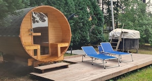 A wooden barrel-shaped structure with a bench and a small table, two blue lounge chairs, and a covered area with a ladder.