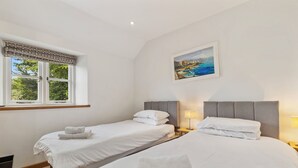 A hotel room with two single beds, a window with a view of greenery, a painting on the wall, and a bedside table with a lamp.