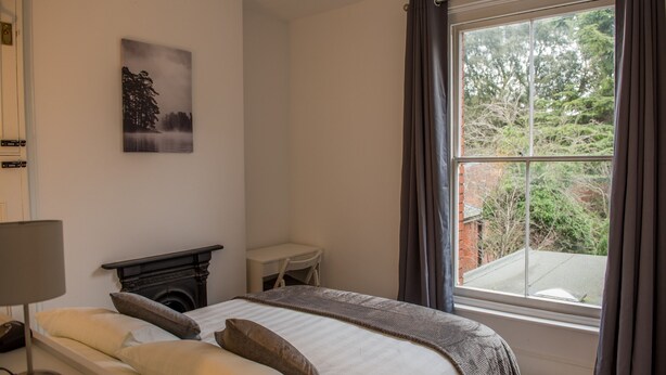 A bedroom with a bed, a fireplace, a desk, and a large window with curtains. - Toothbrush Apartments - Central Ipswich - Fonnereau Rd (Ipswich)