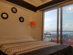 Ein Schlafzimmer mit einem Bett, einem Fenster mit Blick auf eine Stadtlandschaft und einer Wandlampe.