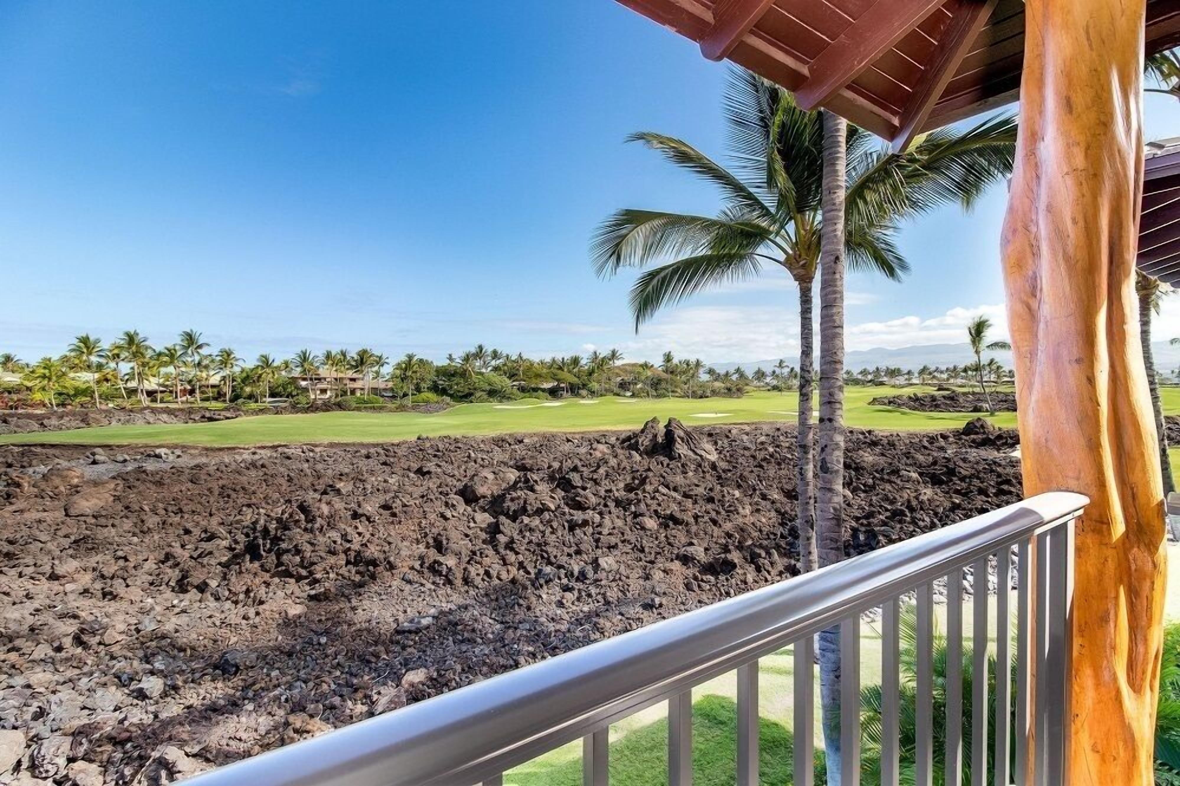 A view from a balcony overlooking a golf course with a lava rock barrier, palm trees, and a clear blue sky.