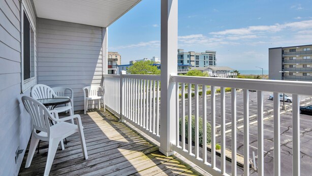 A covered balcony with white railings, two white plastic chairs, and a view of a parking lot and buildings in the background. - Two bedroom, with pool, close to Seacrets! (Ocean City)