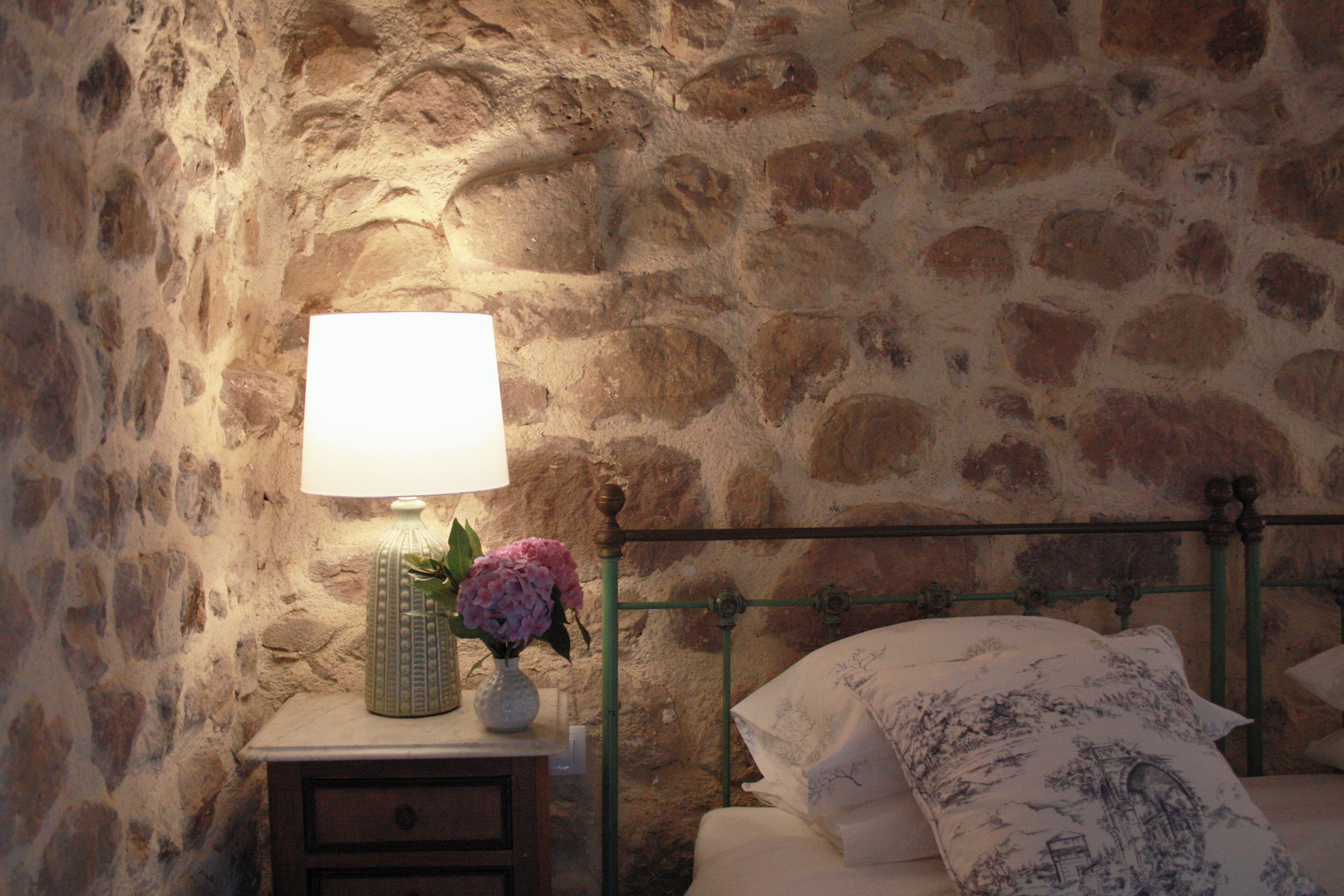 Un mur en pierre, une table de chevet avec une lampe, un vase rempli de fleurs et un lit agrémenté d’oreillers à motifs.