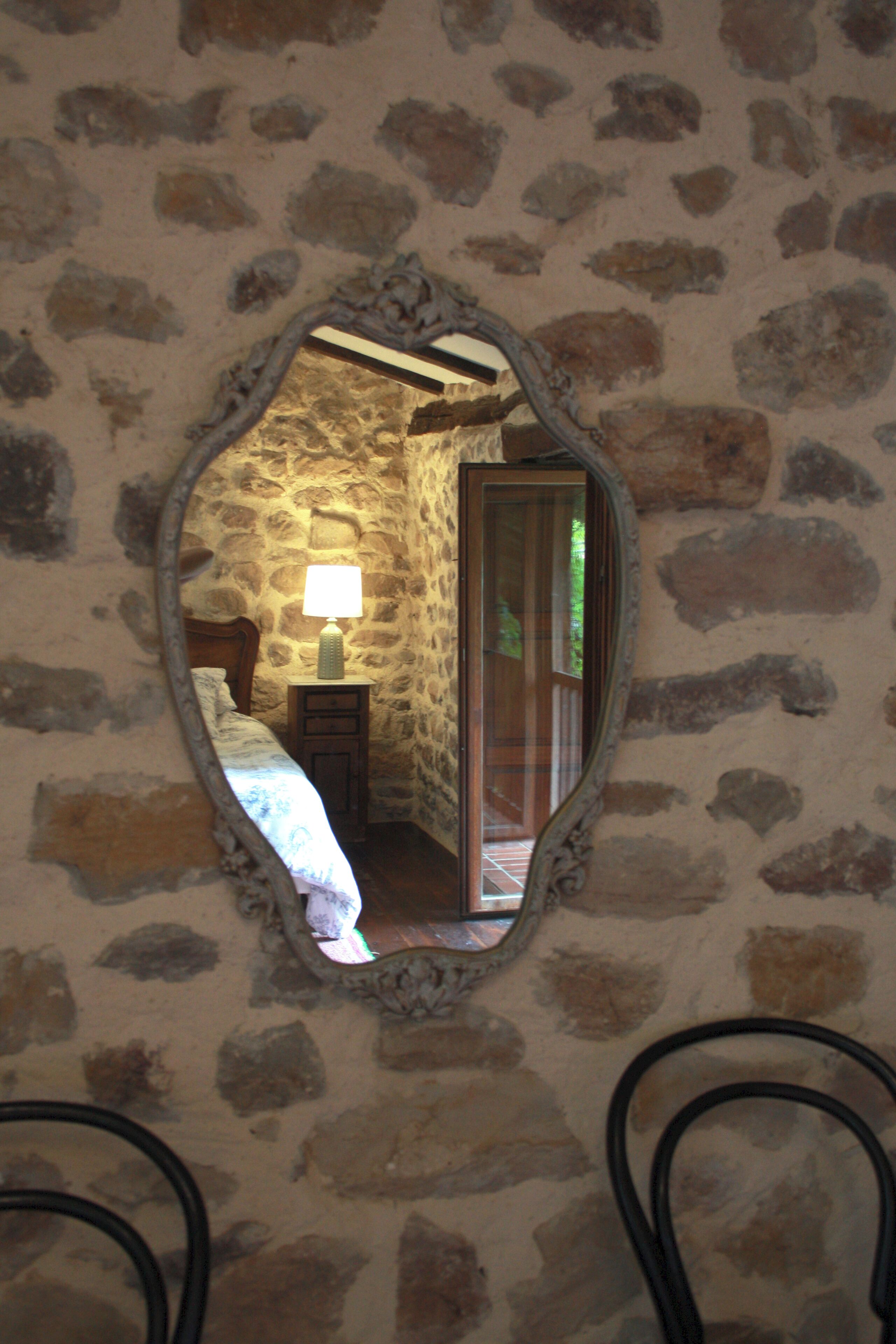 A stone-walled room with a bed, a nightstand with a lamp, and a mirror reflecting the room.