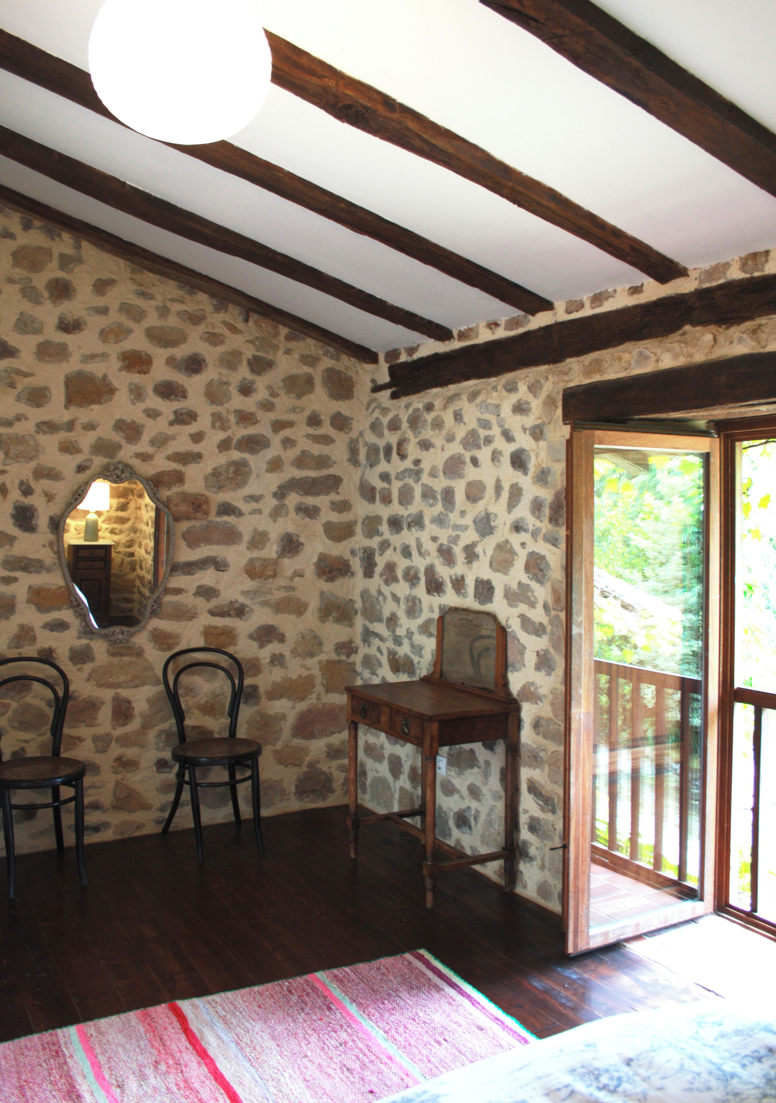 Una habitación con paredes de piedra, vigas de madera, un espejo, dos sillas, una mesa pequeña y una puerta que da a un espacio exterior.