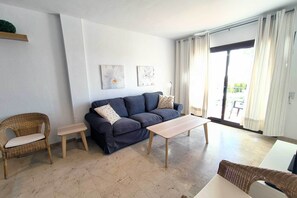 A living room with a dark blue sofa, a small wooden coffee table, wicker chairs, and a sliding glass door leading to an outdoor area.