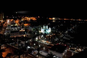 Une vue aérienne nocturne d’une ville côtière, avec un bâtiment imposant en forme de phare, illuminé, qui se dresse au centre, entouré de bâtiments plus petits et de rues.