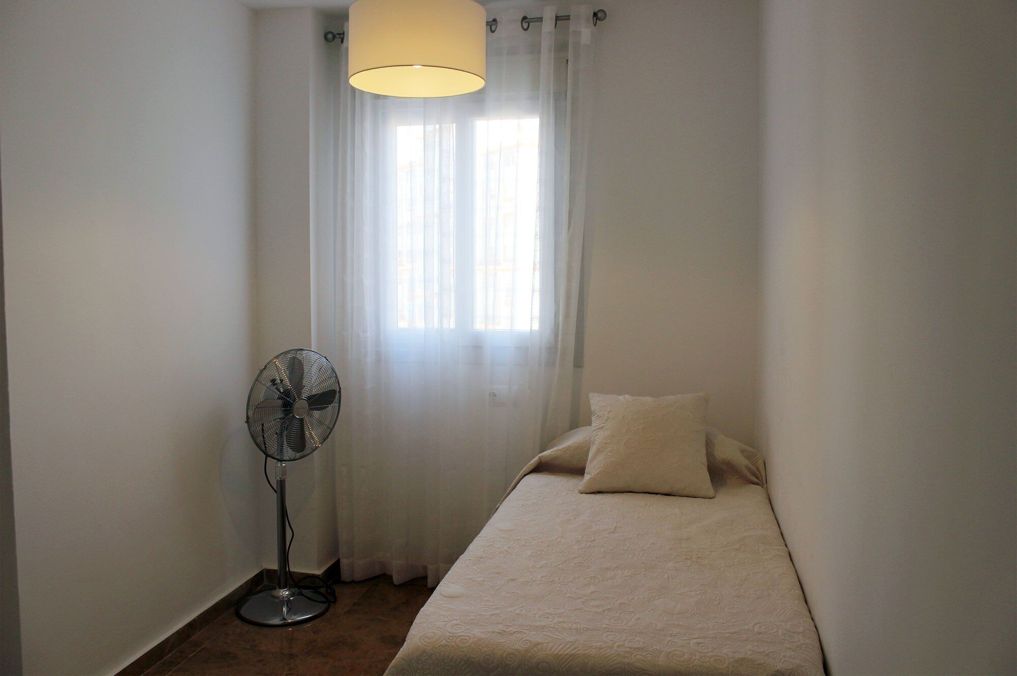 Une petite chambre avec un lit simple, un ventilateur sur pied, une fenêtre avec des rideaux légers et un simple luminaire.