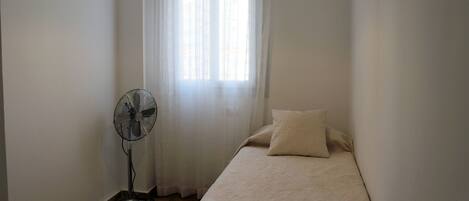 Une petite chambre avec un lit simple, un ventilateur sur pied, une fenêtre avec des rideaux légers et un simple luminaire.