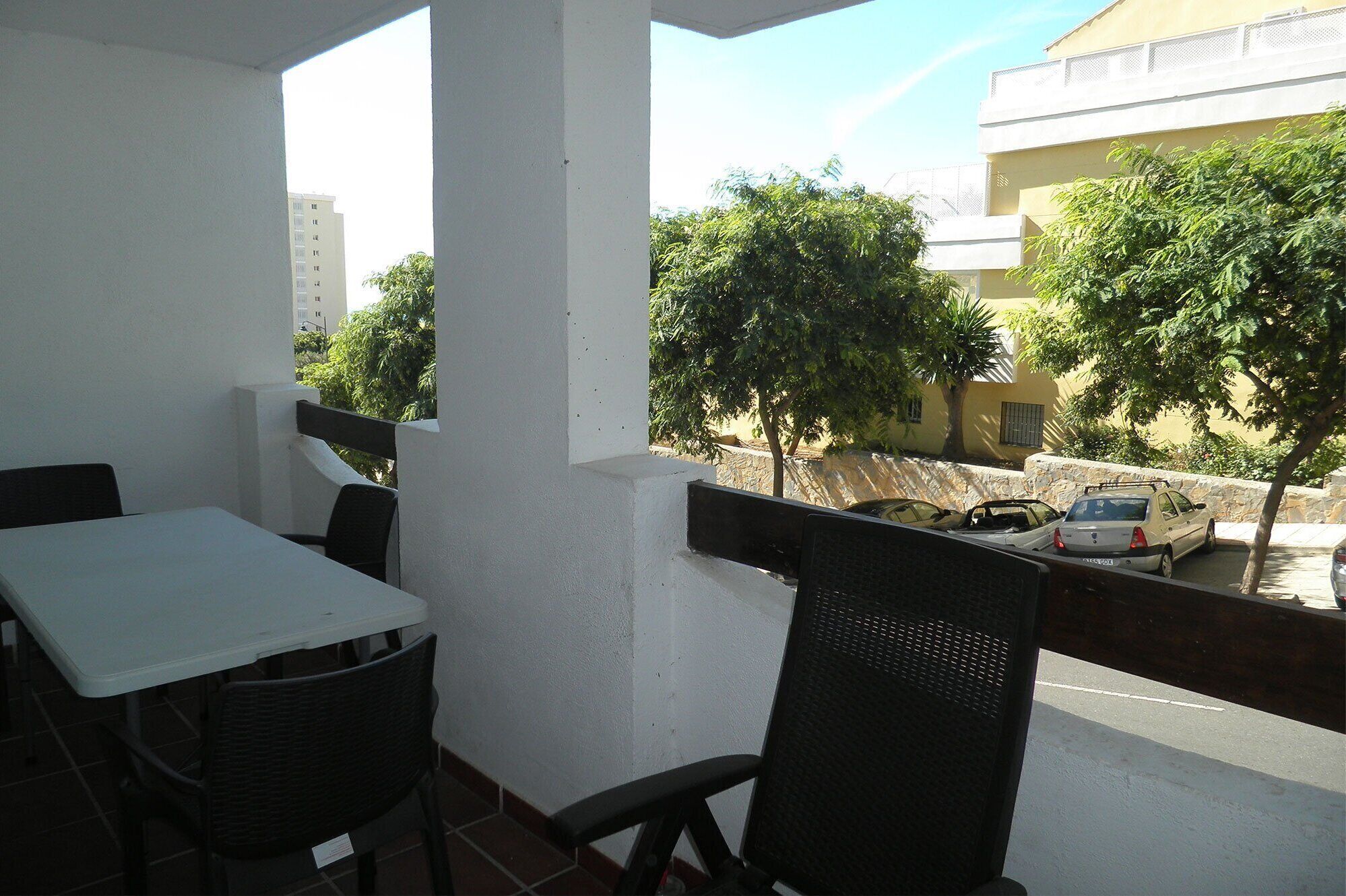 A balcony with a white table and two chairs, overlooking a street with parked cars and trees.