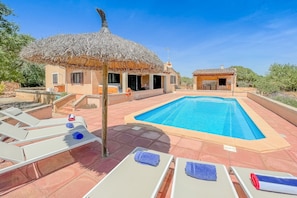 A pool area with lounge chairs, a thatched-roof structure, and a small building with a tiled roof.