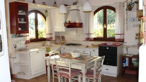Une cuisine traditionnelle avec des meubles blancs, une table à manger recouverte d’une nappe à carreaux et une grande fenêtre agrémentée de rideaux.