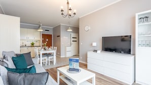 A modern living room with a sofa, coffee table, and TV. There is a kitchen in the background with white cabinets and a dining area.