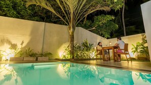An outdoor pool area with a table and chairs, surrounded by plants and a white wall.