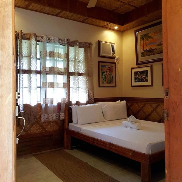 Una habitación pequeña y acogedora con una cama, una silla y un ventanal con cortinas. Hay cuadros enmarcados en la pared y un ventilador de techo.