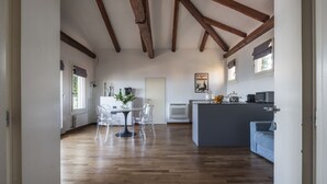Une pièce spacieuse avec des poutres en bois au plafond, un coin repas avec des chaises transparentes, une cuisine dotée d’un plan de travail gris et un canapé bleu.