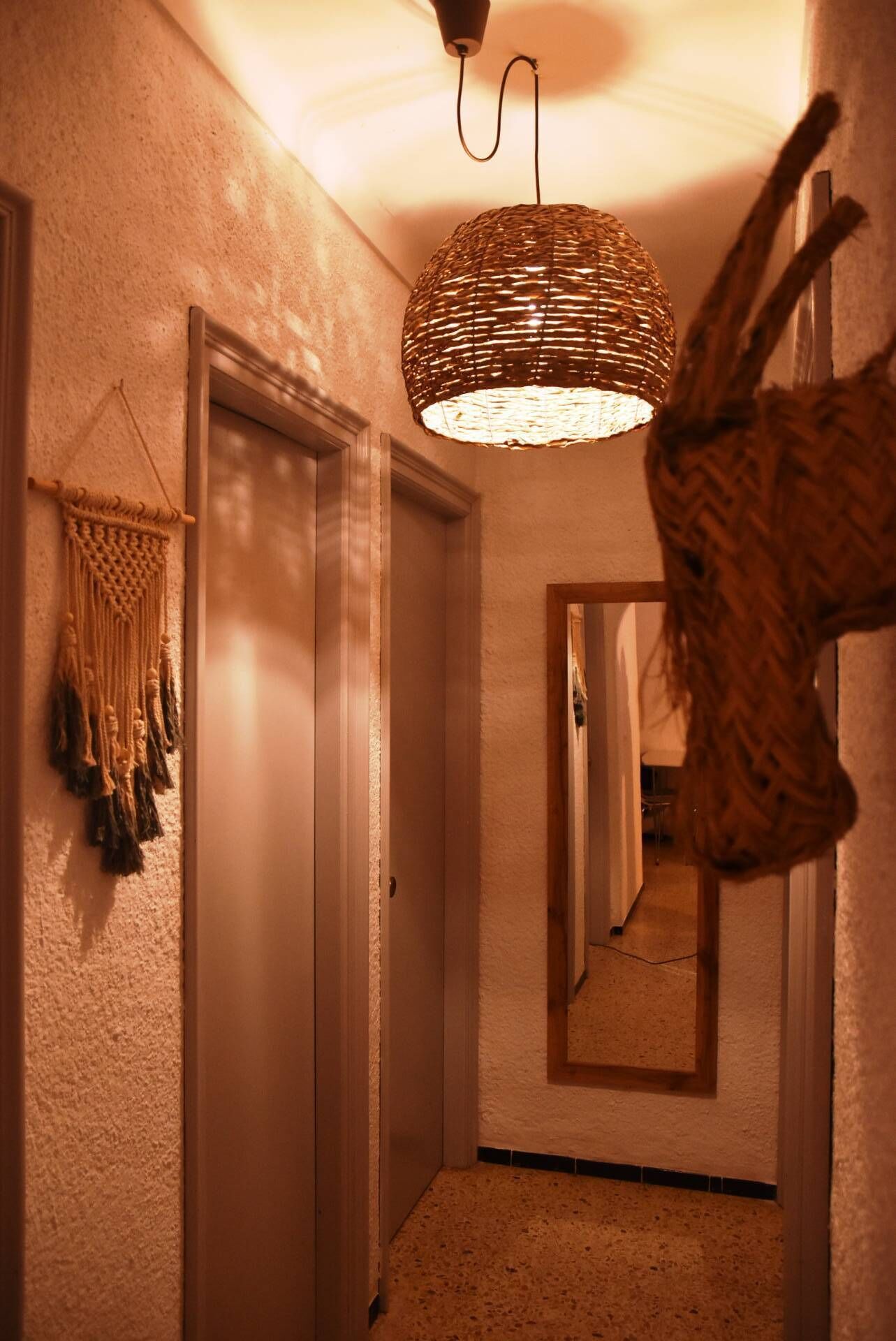 Un couloir agrémenté d’un luminaire tressé, d’une décoration murale en macramé et d’un miroir qui reflète l’espace.